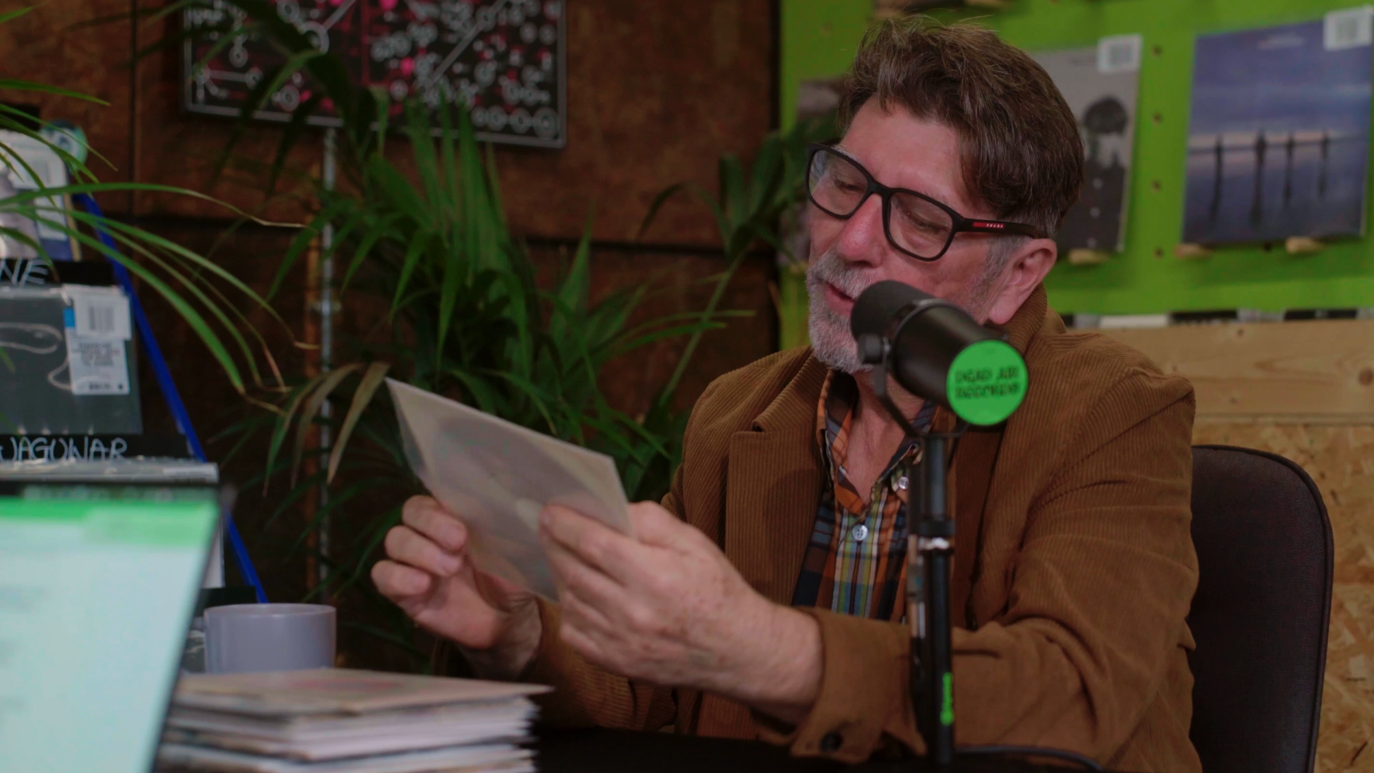 Will Sergeant chatting about vinyl records on the Dead Air Records Podcast, wearing a brown jacket and sat at a desk with Echo and the Bunnymen records on display in the background.